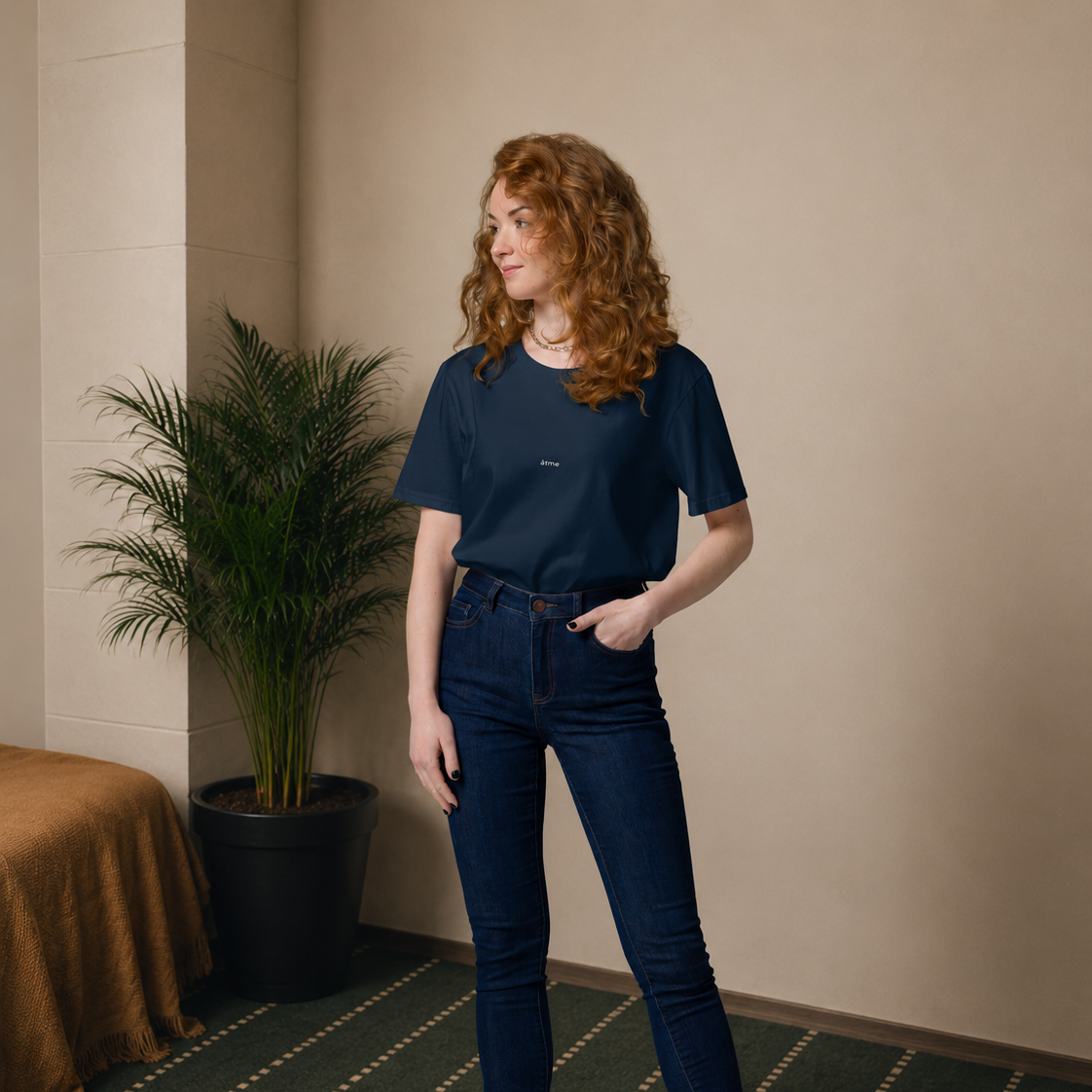 A woman with curly red hair stands in an indoor setting wearing a dark navy blue t-shirt, dark blue skinny jeans, and black ankle boots. She is positioned next to a potted palm tree on a patterned green carpet.