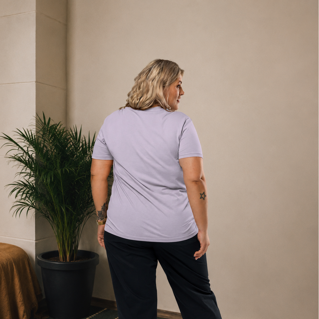 A woman is shown from behind wearing a light lavender short-sleeve t-shirt, dark cropped pants, and white sneakers in an indoor setting with a beige wall, patterned carpet, and a potted plant.